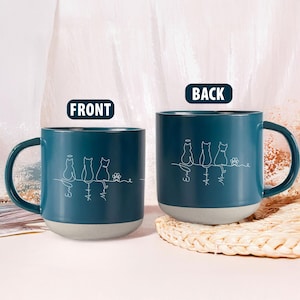 May include: Two teal ceramic mugs with a gray base are displayed. Each mug features a white line drawing of three cats and a flower, with names below. The word "FRONT" and "BACK" are printed on the mugs.