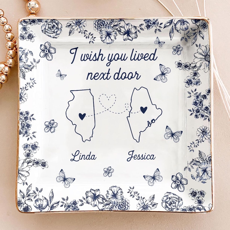 May include: A white ceramic square trinket dish with a gold rim, featuring a blue floral design and the text "I wish you lived next door." The dish includes outlines of two states, connected by a dotted line of hearts, with the names "Linda" and "Jessica" below.