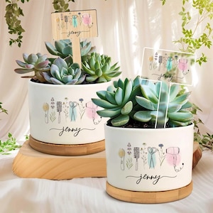 Könnte beinhalten: Zwei weiße Keramik-Sukkulenten-Pflanzgefäße mit Holzsockel. Jedes Pflanzgefäß zeigt ein Design von Friseurwerkzeugen und den Namen "Jenny". Die Sukkulenten sind eine Mischung aus Grün und Blaugrün.