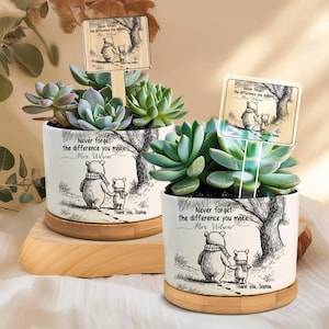 Peut inclure: Deux pots de plantes succulentes en céramique blanche avec une illustration de Winnie l'ourson et le texte "Never forget the difference you make Mrs. Wilson". Chaque pot a une base en bois et une petite pancarte.