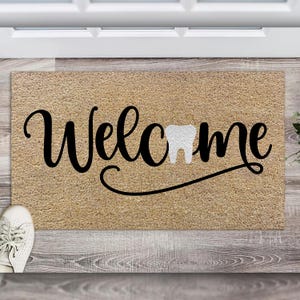 May include: A brown coir doormat with the word "Welcome" in black cursive lettering and a white tooth graphic.