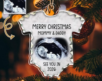 May include: A clear acrylic Christmas ornament shaped like a baby onesie. The ornament features a sonogram image and the text "MERRY CHRISTMAS MOMMY & DADDY SEE YOU IN 2026". The ornament is hanging on a Christmas tree.