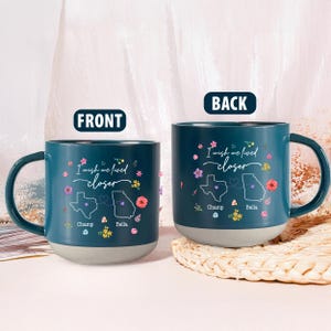 Può includere: Due tazze in ceramica verde acqua con base grigia. Le tazze presentano la scritta "I wish we lived closer" e i contorni degli stati di Texas e Georgia, con le città di Champ e Bella. Accenti floreali decorano le tazze.