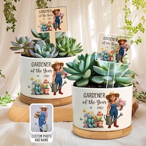 Op de afbeelding: Twee witte keramische plantenbakken met vetplanten, elk met een "Tuinman van het Jaar" ontwerp met een cartoon illustratie van een tuinman. De plantenbakken staan op houten bases en bevatten een houten bord.