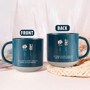 Pode incluir: Duas canecas de cerâmica azul-petróleo com base cinza e alça curva. Cada caneca apresenta um design floral com os nomes "Bella" e "Helen" e o texto "IT TAKES A LONG TIME TO GROW AN OLD FRIEND."