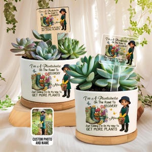 May include: Two white ceramic planters with succulents, each featuring the text "I'm A Plantaholic On The Road To Recovery" and a cartoon illustration. One planter includes a small photo. The planters sit on wooden bases.