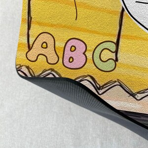 May include: A yellow and white yoga mat with a black non-slip surface. The mat features a colourful illustration of the letters "ABC" with a flower and a line drawing of a face.