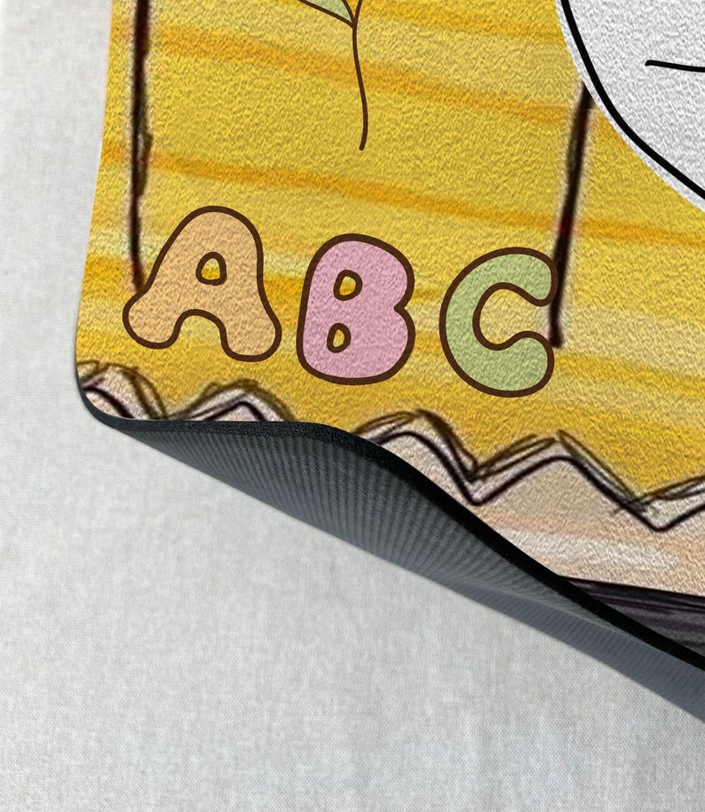 May include: A yellow and white yoga mat with a black non-slip surface. The mat features a colourful illustration of the letters "ABC" with a flower and a line drawing of a face.