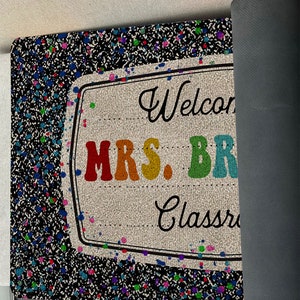 May include: A black welcome mat with a rainbow-colored sign that says "Welcome to Mrs. Brown's Classroom". The sign is surrounded by colorful paint splatters.