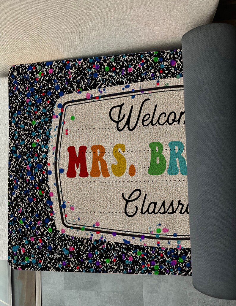 May include: A black welcome mat with a rainbow-colored sign that says "Welcome to Mrs. Brown's Classroom". The sign is surrounded by colorful paint splatters.