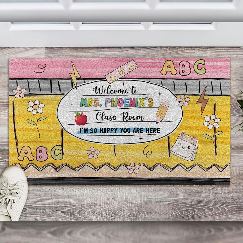 May include: A yellow and pink welcome mat with the text "Welcome to Mrs. Phoenix's Class Room I'm so happy you are here" and the letters "ABC" on either side.