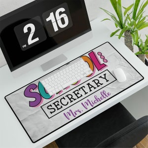 May include: A white desk mat with a crumpled paper texture. The text "School Secretary" is in a black rectangle with a white border. The word "School" is in colorful letters with a rainbow effect. The text "Mrs. Michelle" is in purple below the rectangle.