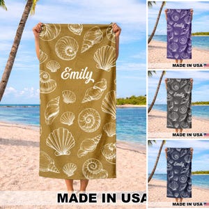 May include: A beach towel with a tan background and a white seashell pattern. The name "Emily" is printed in white script. The towel is held up on a beach with the text "MADE IN USA" at the bottom.