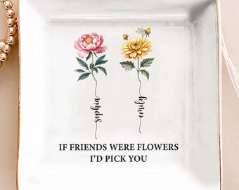 Op de afbeelding: Een witte vierkante keramische sieradenschaal met een gouden rand. De schaal heeft twee bloemenillustraties, een roze pioenroos en een gele chrysant, met de namen "Sophia" en "Emily" eronder geschreven. De tekst "IF FRIENDS WERE FLOWERS I'D PICK YOU" staat onderaan.