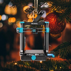 May include: A black and teal 3D printer ornament with the name "MICHAEL" printed on it. The ornament is hanging from a string and is set against a blurred background of Christmas lights and a red ornament.