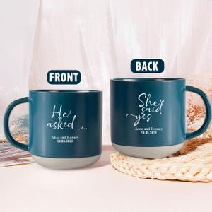 Può includere: Due tazze in ceramica verde acqua con testo bianco. Una tazza recita "He asked..." e l'altra "She said yes". Entrambe le tazze riportano i nomi "Anna and Ronney" e la data "20.08.2023". Le tazze hanno basi grigie.