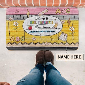 May include: A yellow and pink welcome mat with the text "Welcome to Mrs. Phoenix's Class Room I'm so happy you are here" and the letters "ABC" on it. The mat has a cartoon style with flowers, lightning bolts, and a calendar.