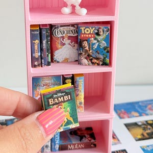 Peut inclure: Une bibliothèque miniature rose à trois étagères avec une figurine blanche de Mickey Mouse sur l'étagère supérieure et divers petits livres de films Disney. Une main tient un petit livre Bambi devant l'étagère, avec d'autres couvertures de livres.