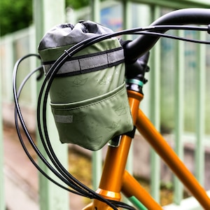 Puede incluir: Una bolsa de manillar verde con un patrón de barcos de papel unida a una bicicleta naranja brillante con manillar y cables negros. La bolsa tiene una banda reflectante y el texto "VALENTINO DIVISION" es visible.