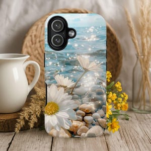 May include: A phone case featuring a serene beach scene with white daisies on a pebble beach. The background shows sparkling blue water and a clear sky. The case has a glossy finish and a camera cutout.