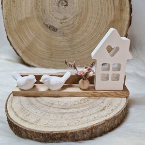 Könnte beinhalten: Zwei weiße Keramikvögel sitzen auf einem Holzregal mit einem weißen Keramikhaus mit einem Herz-Ausschnitt und einem kleinen Blumenstrauß in einem Topf. Das Regal steht auf einer Holzscheibe.