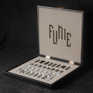 May include: A black wooden box with a grey felt interior. The box is open and contains a set of silver chess pieces. The box has the word "FUME" cut out of the felt.