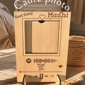 Peut inclure: Un cadre photo en bois avec une découpe pour une photo. Le cadre a un logo Spotify, un code-barres et le texte "ROSE BRUNO MARS APT." Il a également le texte "Best Song" et "Madhi" avec un symbole de cœur.