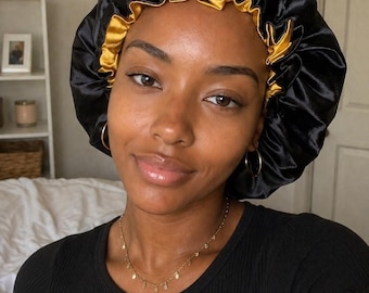 May include: A black satin bonnet with a gold interior. The bonnet is worn by a person with a gold necklace and hoop earrings. The person is wearing a black shirt. The background includes a white shelf and a bed.