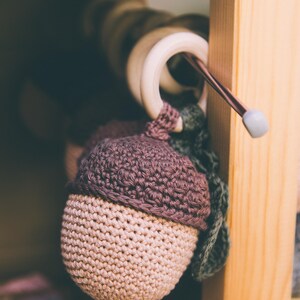 Könnte beinhalten: Ein handgehäkeltes Eichelspielzeug in Beige und Lila, mit einem grünen Blattdetail. Die Eichel hängt an einem Holzring, was auf ein Baby- oder Kleinkindspielzeug hindeutet. Der Hintergrund ist eine Holzkonstruktion.