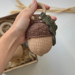 Könnte beinhalten: Eine gehäkelte Eichel-Rassel mit brauner Kappe und beigefarbenem Körper. Die Rassel hat einen daran befestigten Holzring und ein grünes gehäkeltes Blattdetail.