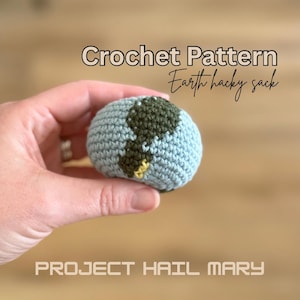 Puede incluir: Un hacky sack de la Tierra tejido a ganchillo, con océanos azules y continentes verdes. Se muestra el texto "Crochet Pattern Earth hacky sack" y "PROJECT HAIL MARY". El hacky sack se sostiene en una mano.