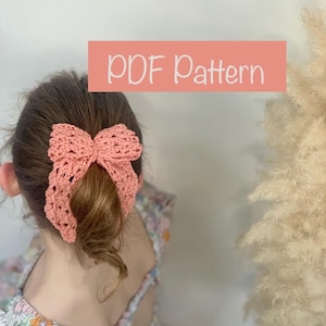 Könnte beinhalten: Ein gehäkeltes, korallenfarbenes Schleifen-Stirnband wird im Haar getragen. Der Text "PDF Pattern" wird auf einem rosa rechteckigen Banner angezeigt. Die Person trägt ein Kleid mit Blumenmuster.