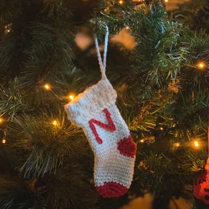 Puede incluir: Un pequeño adorno de calcetín navideño de ganchillo en color blanco roto, con una letra N roja bordada en la parte delantera, un talón rojo y una puntera roja. El calcetín incluye un lazo de hilo blanco para colgarlo.