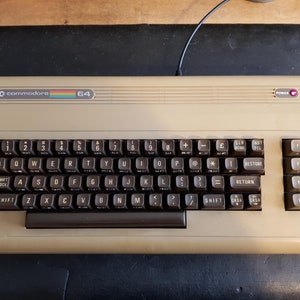 May include: A beige Commodore 64 computer with a black keyboard and a rainbow logo. The keyboard has keys labelled with letters, numbers, and symbols. There is a red power light on the top right corner of the computer.
