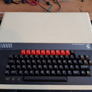 May include: A beige and black BBC Microcomputer with a red and black keyboard. The keyboard has function keys labelled F1 through F10. The computer has a cassette motor and a cap lock.