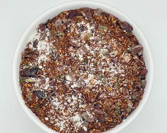 May include: A white ceramic bowl filled with a mix of loose-leaf tea. The tea blend features a reddish-brown base with visible pieces of cocoa nibs, green herbs, and white powder. The tea is presented against a plain white background.