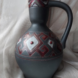 May include: A black ceramic vase with a red and white geometric design. The vase has a wide base and a narrow neck with a handle.