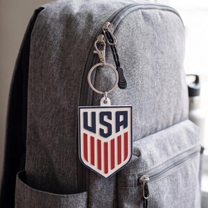 May include: A grey fabric backpack featuring a shield-shaped keychain attached to the zip. The keychain is white with dark blue 'USA' text at the top and vertical red stripes below, all encased in a dark blue border with a silver metal ring.