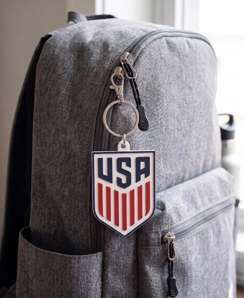 May include: A grey fabric backpack featuring a shield-shaped keychain attached to the zip. The keychain is white with dark blue 'USA' text at the top and vertical red stripes below, all encased in a dark blue border with a silver metal ring.