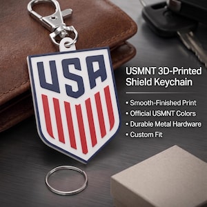 May include: A shield-shaped keyring featuring the letters USA in dark blue above red vertical stripes on a white background. The keyring includes a silver metal lobster clasp and a separate circular ring. Text on the image reads USMNT 3D-Printed Shield Keychain, Smooth-Finished Print, Official USMNT Colors, Durable Metal Hardware, Custom Fit, and Show Your Pride on the Go.