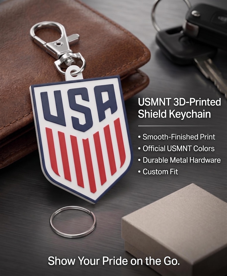 May include: A shield-shaped keyring featuring the letters USA in dark blue above red vertical stripes on a white background. The keyring includes a silver metal lobster clasp and a separate circular ring. Text on the image reads USMNT 3D-Printed Shield Keychain, Smooth-Finished Print, Official USMNT Colors, Durable Metal Hardware, Custom Fit, and Show Your Pride on the Go.