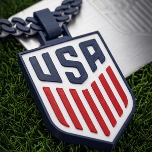 May include: A navy blue and white USA pendant necklace with a matching chain. The pendant features a shield design with red stripes and the letters "USA" in navy blue. The necklace is displayed on a bed of green grass.