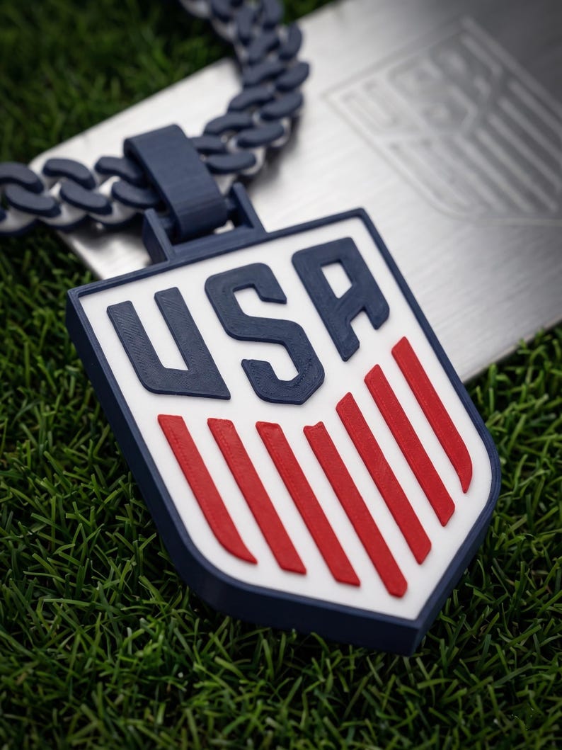 May include: A navy blue and white USA pendant necklace with a matching chain. The pendant features a shield design with red stripes and the letters "USA" in navy blue. The necklace is displayed on a bed of green grass.