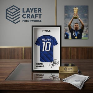 Peut inclure: Un cadre 3D pr&eacute;sentant un maillot de football bleu de la France avec le nom MBAPPE et le num&eacute;ro 10 en blanc. L'affichage inclut le texte FRANCE, KYLIAN MBAPPE et une signature. Une plaque dor&eacute;e indique LAYER CRAFT PREMIUM COLLECTOR'S SERIES.