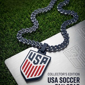 May include: A USA Soccer Fan Gear necklace with a shield pendant. The pendant features the letters "USA" in blue and red stripes on a white background. The chain is a dark gray and white link design. The text "COLLECTOR'S EDITION USA SOCCER FAN GEAR" is also visible.