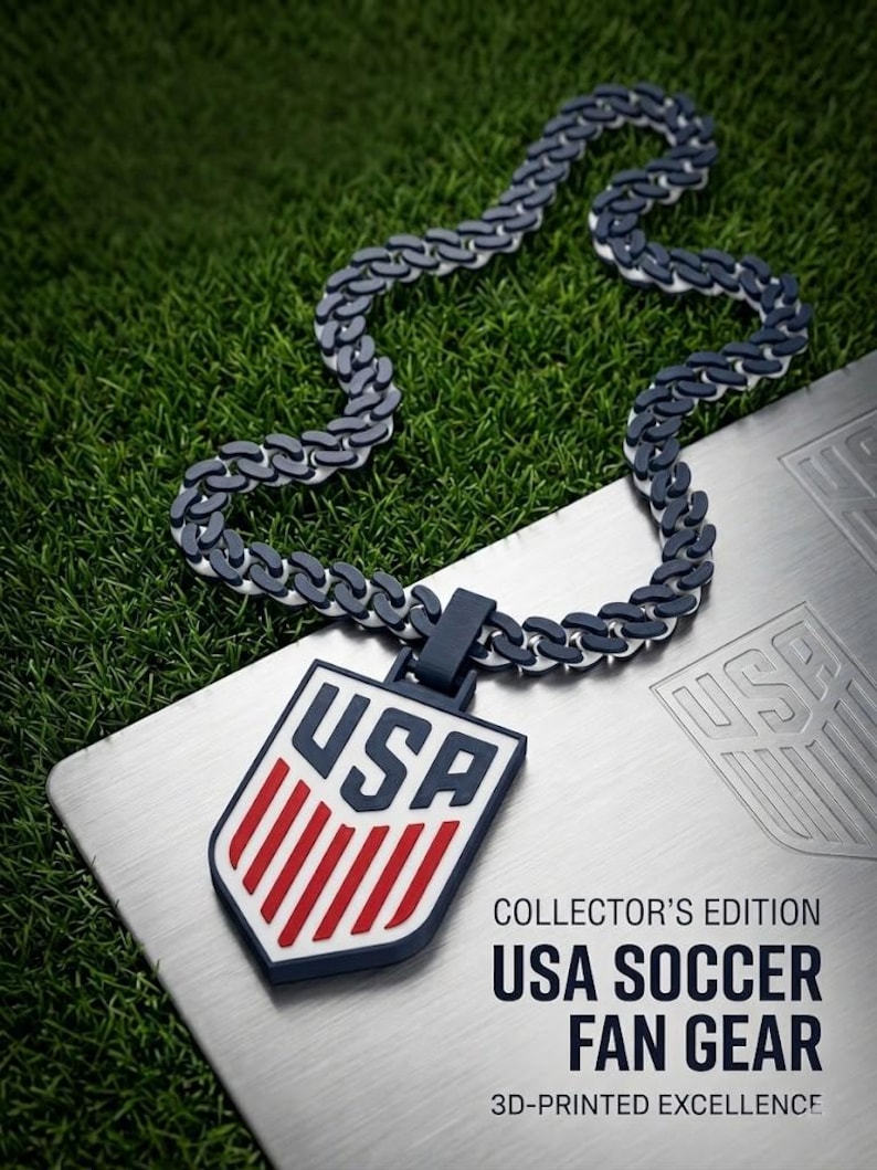 May include: A USA Soccer Fan Gear necklace with a shield pendant. The pendant features the letters "USA" in blue and red stripes on a white background. The chain is a dark gray and white link design. The text "COLLECTOR'S EDITION USA SOCCER FAN GEAR" is also visible.