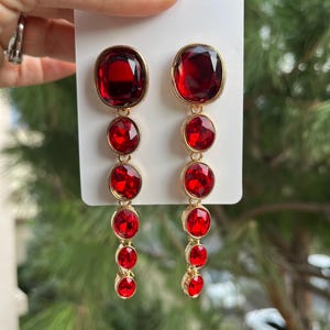 Op de afbeelding: Een paar bungelende oorbellen met een goudkleurige zetting. Elke oorbel heeft een grote, ovale, dieprode edelsteen aan de bovenkant, gevolgd door drie kleinere, ronde rode edelstenen. De oorbellen worden tegen een witte achtergrond gehouden.