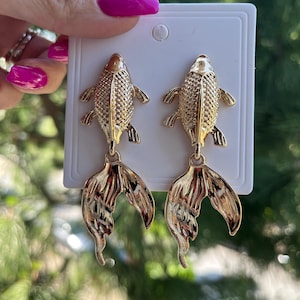 Può includere: Orecchini a forma di pesce color oro. Ogni orecchino presenta un corpo di pesce dettagliato con squame strutturate e pinne caudali fluenti. Gli orecchini sono esposti su una carta bianca, con uno sfondo verde sfocato.
