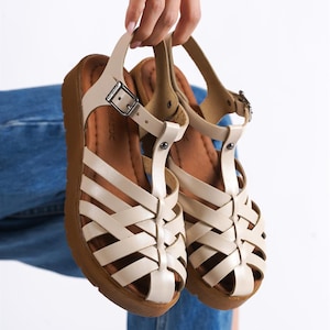 May include: A pair of cream-colored sandals with a woven design and a brown sole. The sandals have a strap that buckles at the ankle. The shoes are held up by a person.