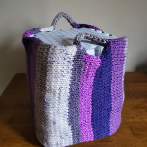 Può includere: Un cesto di stoccaggio a maglia con un motivo a righe in tonalità di grigio, viola e rosa. Il cesto ha due manici ed è realizzato con un filato spesso.
